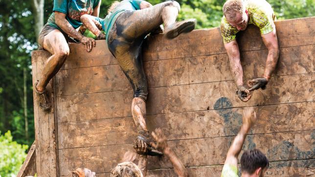 A team helping one another over a wall in a muddy obstacle course - teamwork crisis